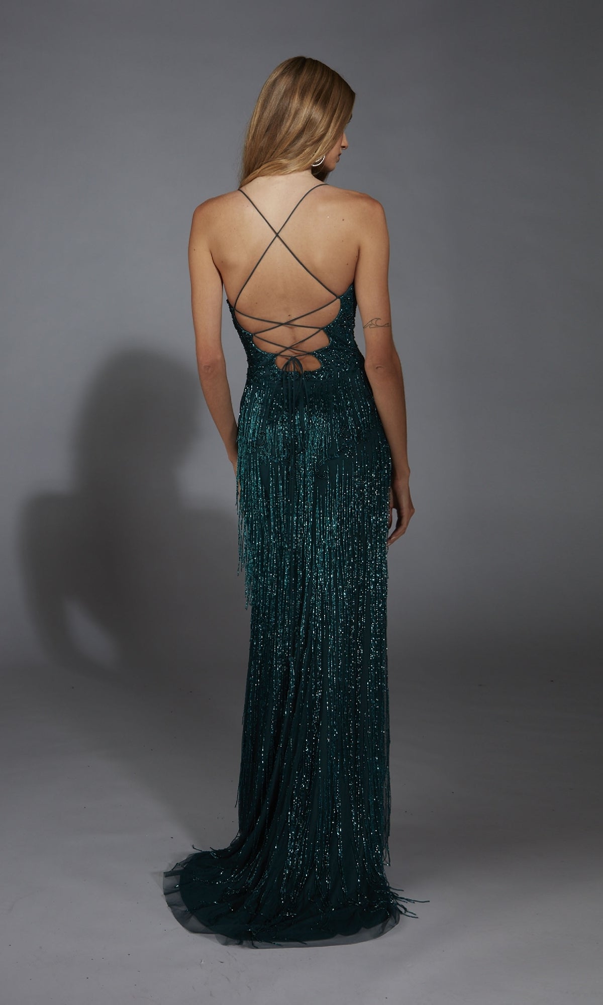 A woman with long hair stands facing away in the Alyce 61788, a sparkly dark teal prom dress with beaded fringe, a floor-length fringed skirt, and an open back with crisscross straps.